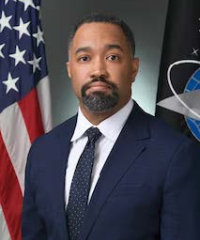 A headshot of Russell L. Hudgins the 2nd in a navy suit and tie infront of an United States flag and Unitied States Space Force flag